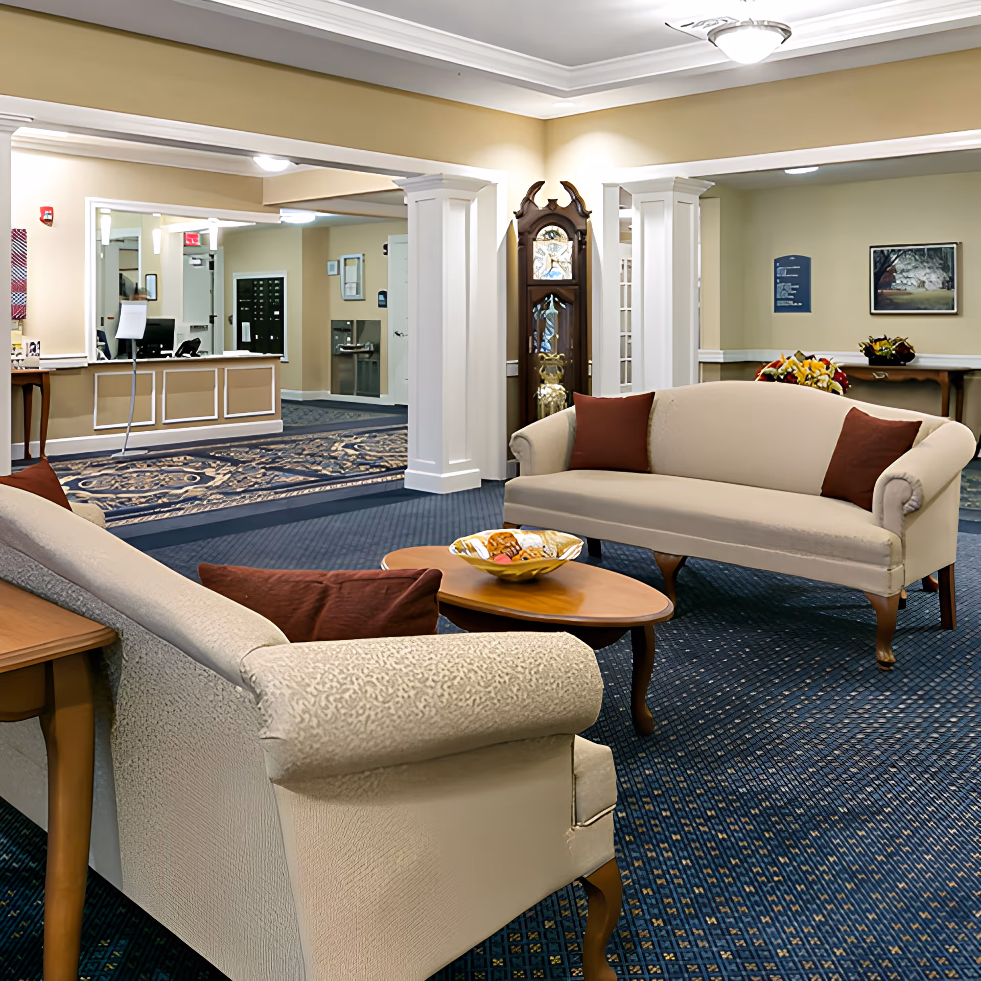 A cozy assisted living facility lobby with two beige upholstered sofas with red cushions facing each other across a wooden coffee table holding a decorative bowl. The room features blue patterned carpet, cream-colored walls, white columns, a grandfather clock, and a reception desk in the background.