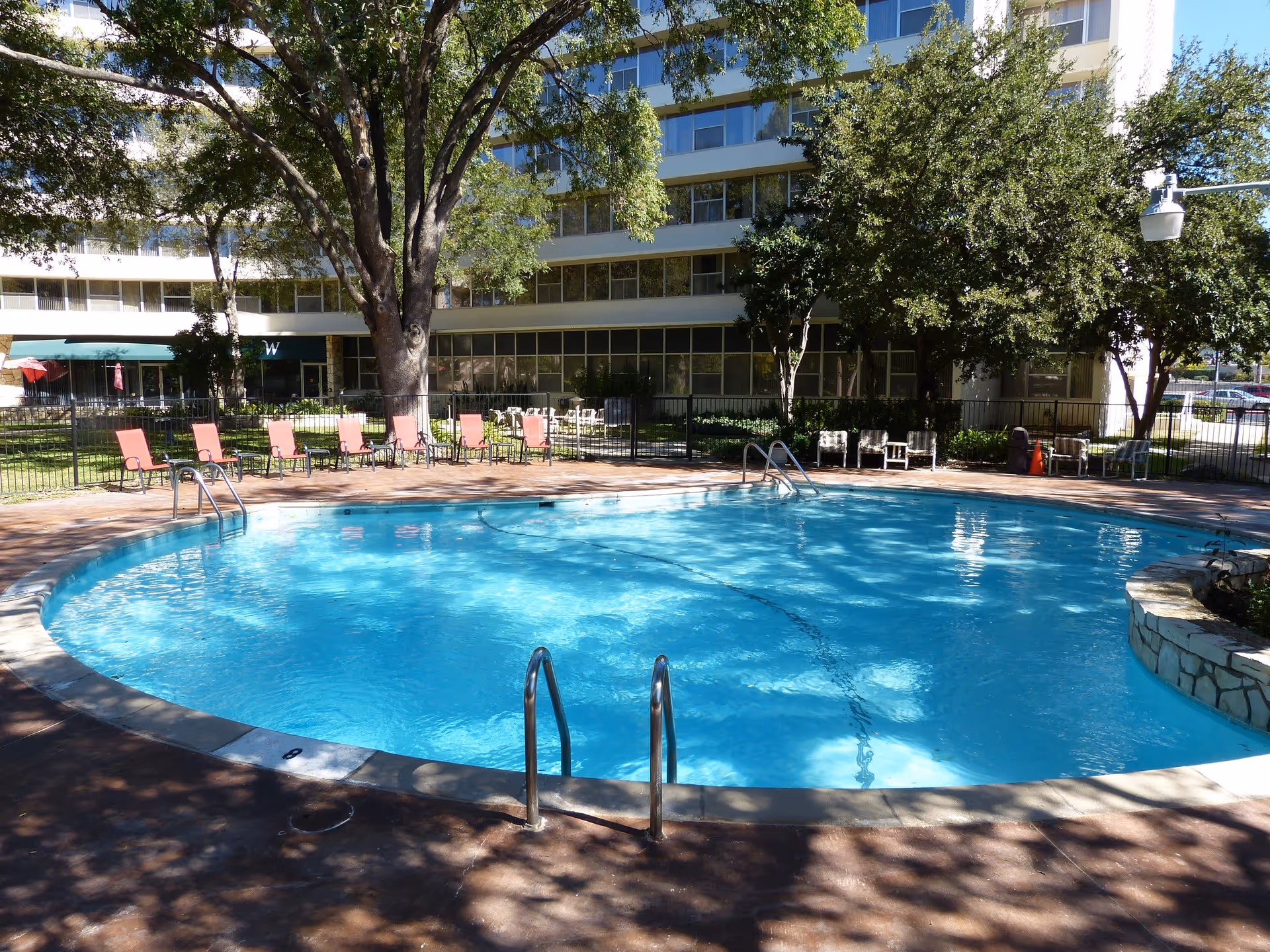 Outdoor swimming pool with clear blue water surrounded by a concrete deck and several red lounge chairs. The pool area is fenced and shaded by large trees. In the background, there is a multi-story building with many windows and some outdoor seating areas.