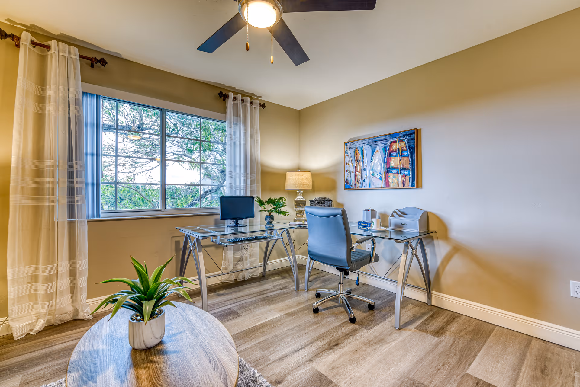 A bright office space with a glass desk, a blue office chair, a computer monitor, a lamp, and decorative items. There is a large window with sheer curtains letting in natural light and a ceiling fan above. A small round wooden table with a potted plant is in the foreground, and a colorful painting hangs on the beige wall.