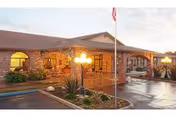 Covered brick entrance of a single-story care facility with arched windows, a flagpole, lamp posts, and a wet driveway.