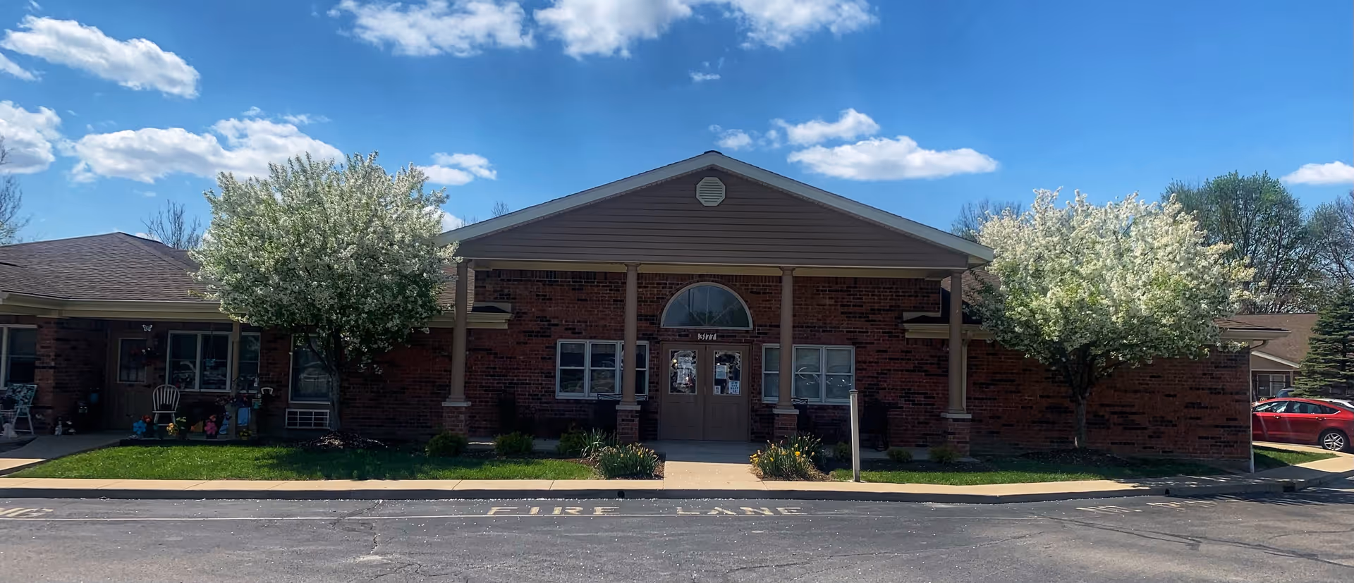 Front exterior view of a single-story brick building with a peaked roof and two white blossoming trees on either side of the entrance. The entrance has double doors with a semicircular window above them. There is a sidewalk leading to the entrance and a parking area in front with 'FIRE LANE' painted on the pavement. The sky is blue with scattered clouds.