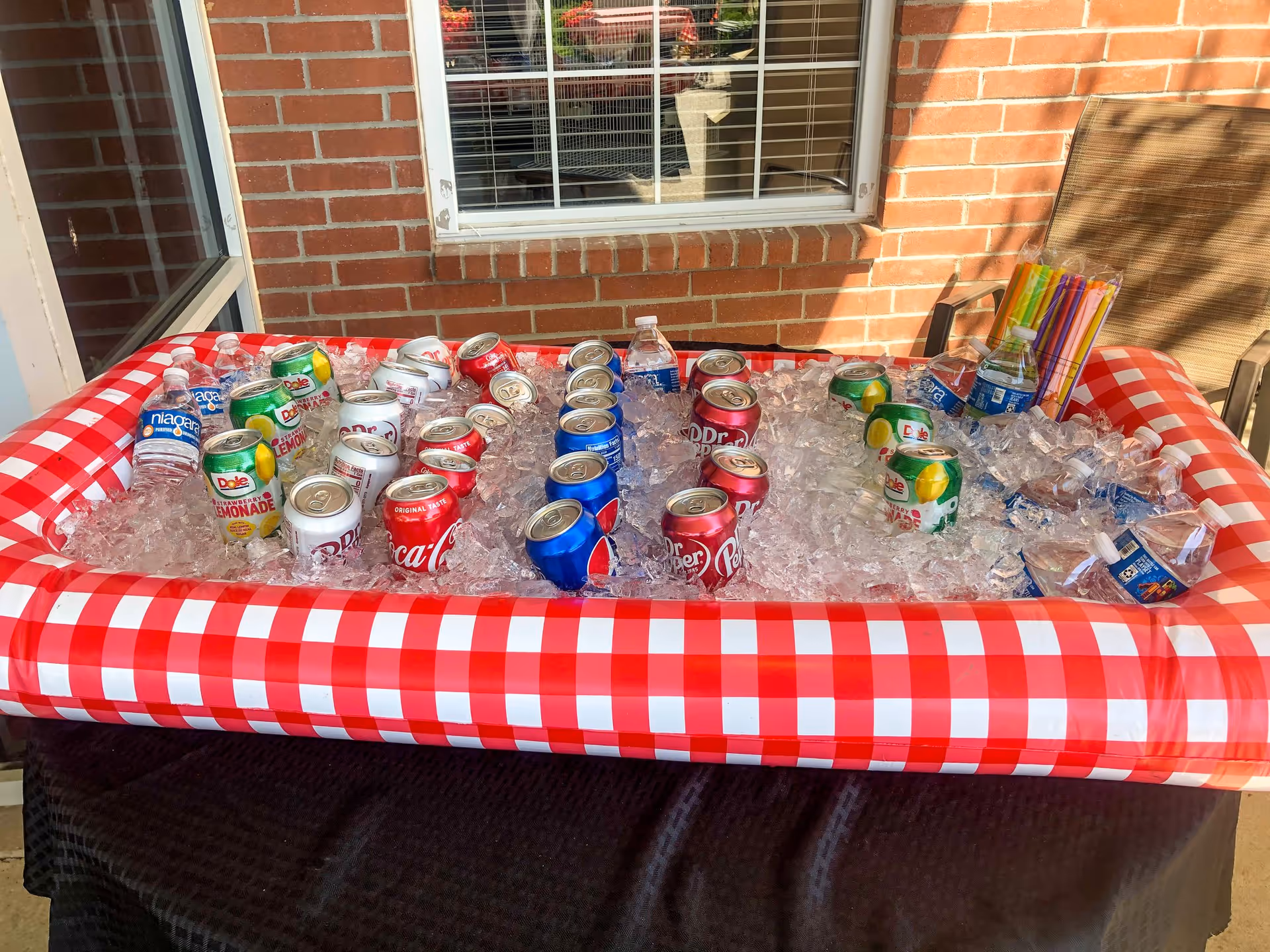 An inflatable red-and-white checkered cooler on a table filled with ice holding assorted canned sodas and bottled water outside against a brick wall and window.