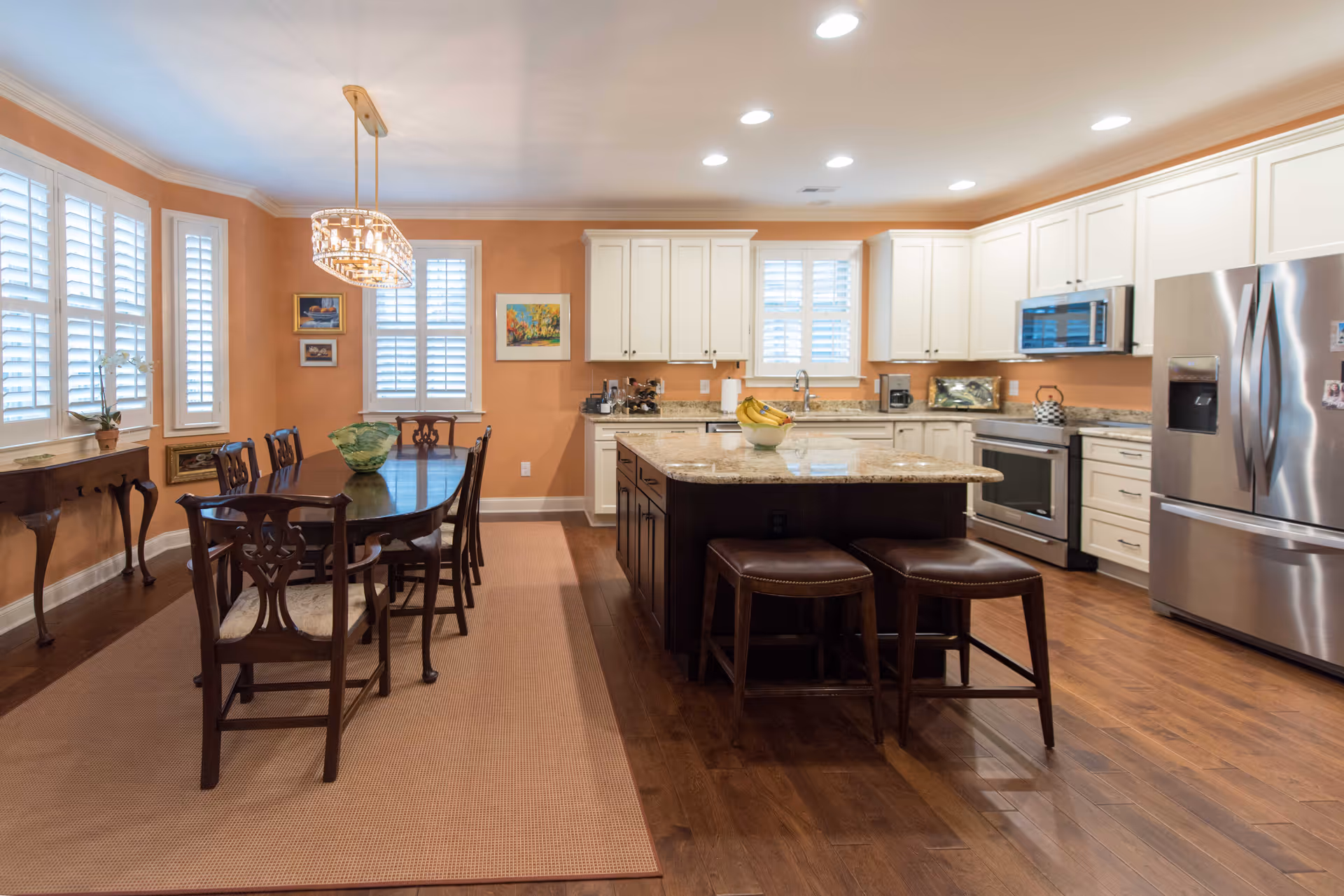 A spacious kitchen and dining area with wooden floors and peach-colored walls. The kitchen features white cabinets, stainless steel appliances including a refrigerator, oven, and microwave, and a large granite island with two brown stools. The dining area has a dark wooden table with six matching chairs and a decorative chandelier above. Windows with white shutters allow natural light into the room, and there are framed pictures on the walls.