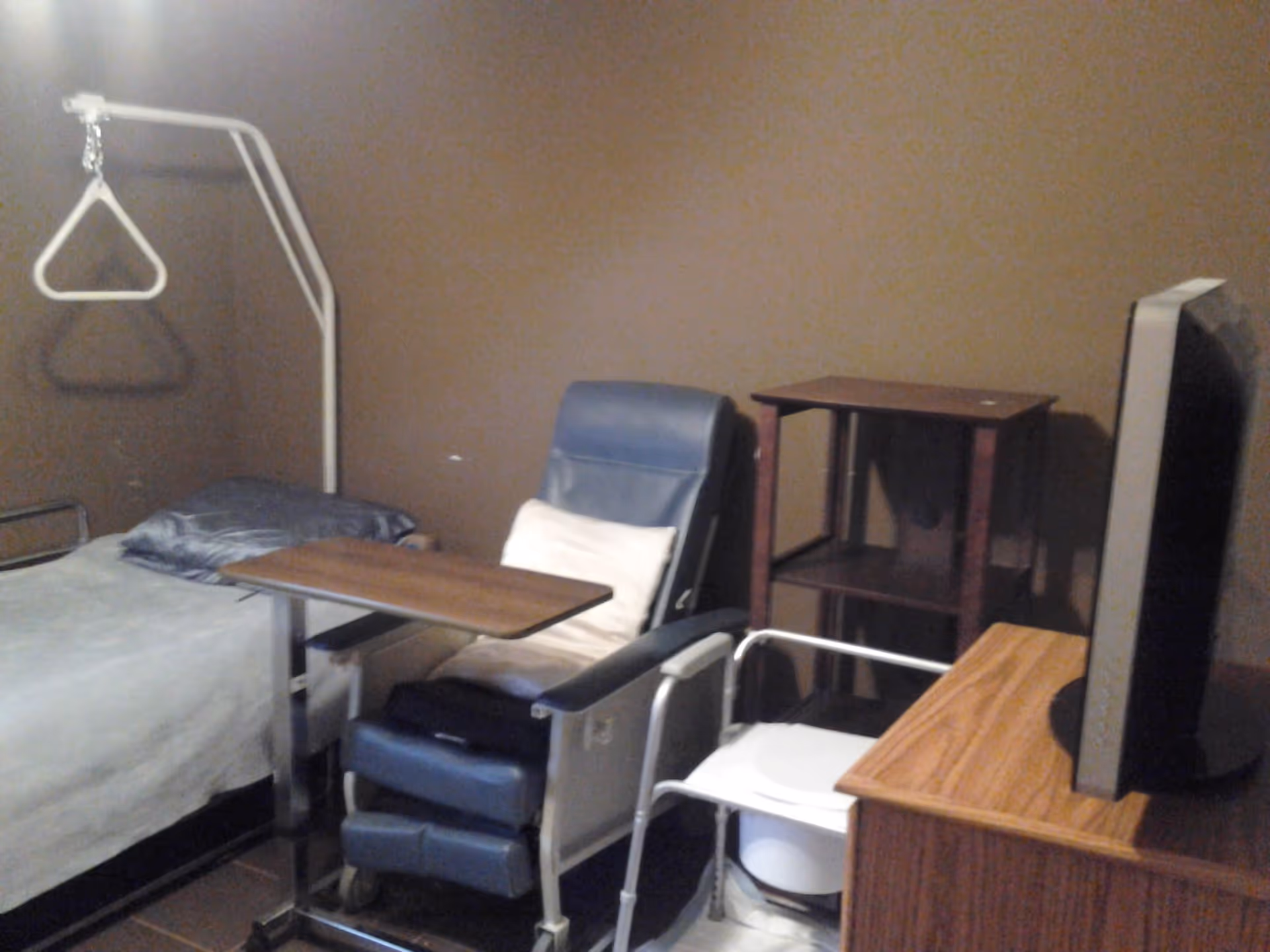 A small bedroom with a hospital-style bed, overbed table, blue recliner, TV and bedside shelving against a brown wall.