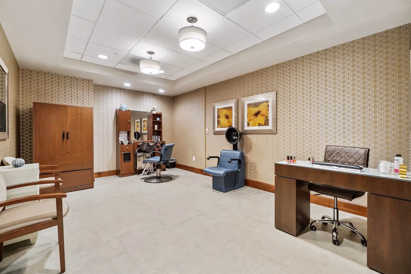 Well-lit salon/styling room with a barber chair, hooded hair dryer, manicure desk, seating, and framed floral artwork.