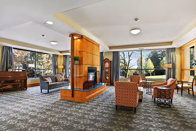 A spacious and well-lit living room area with large windows showing a view of trees and greenery outside. The room features a central wooden fireplace with glass panels, surrounded by various seating options including patterned armchairs, a gray sofa, and side tables. There is a piano against one wall and a tall grandfather clock near the windows. The carpet has a floral pattern, and the room has ceiling lights and curtains framing the windows.