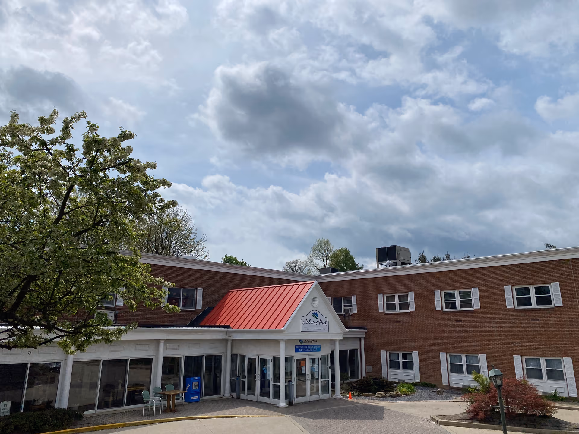 Exterior view of Arbutus Park Retirement Community building with a red roof over the entrance, brick walls, multiple windows, a tree with green leaves on the left, and a cloudy sky overhead.