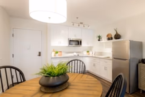 Round wooden dining table with a potted plant in front of a compact white kitchen with stainless steel appliances.