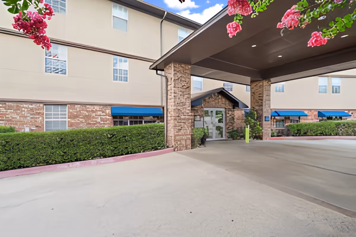 Entrance area of Morada Meadow Lakes facility showing a covered drop-off zone with stone pillars, beige building exterior with multiple windows, blue awnings, green bushes, and pink flowers hanging from above.