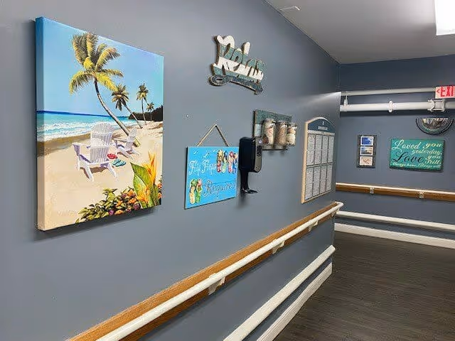 A hallway with gray walls decorated with beach-themed artwork and signs, including a painting of a beach with palm trees and chairs, a sign that says 'Relax', and other decorative plaques. The hallway has wooden handrails on both sides and a dark wood floor.