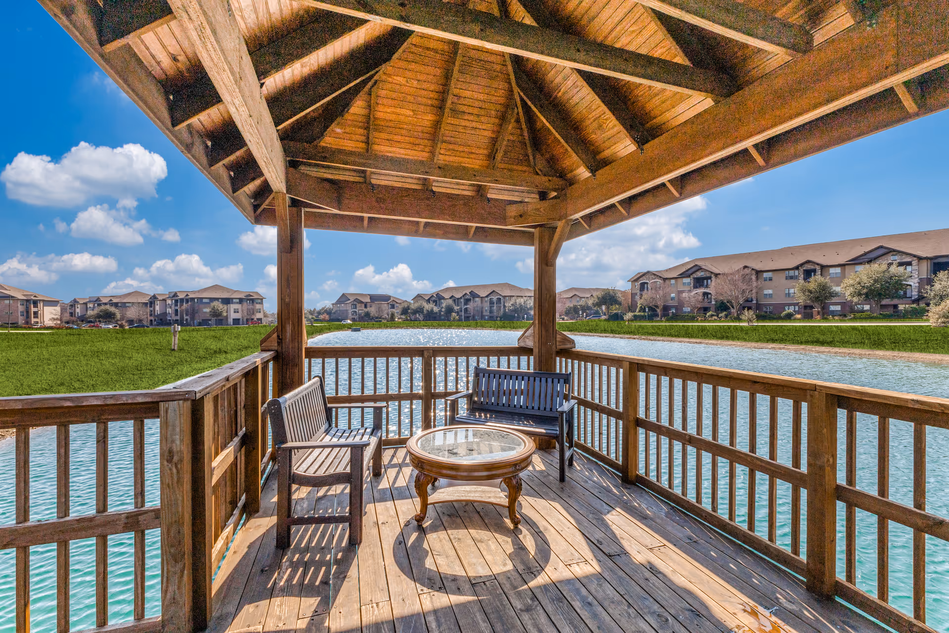 Wooden gazebo with a round glass-top table and two benches overlooking a large pond, with multi-story residential buildings and green lawns in the background under a blue sky with scattered clouds.
