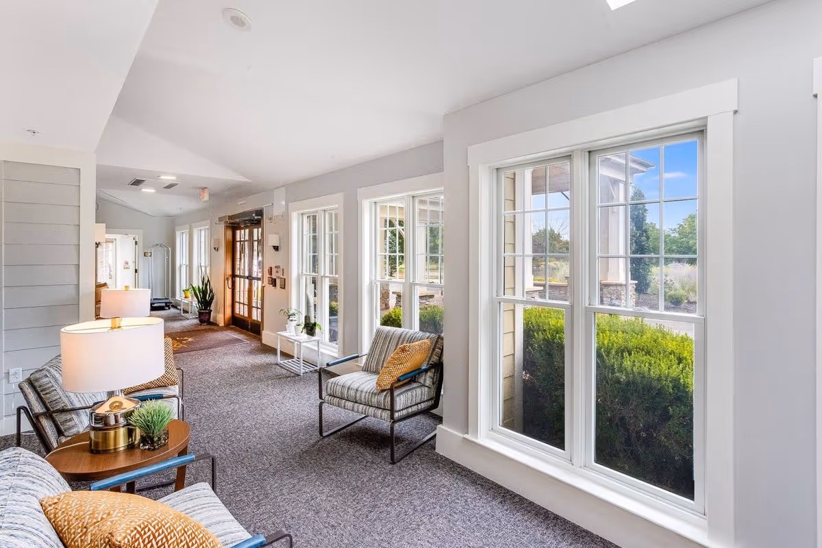 A bright and airy interior hallway with large windows on the right side showing green bushes and a clear blue sky outside. The hallway features comfortable seating with striped chairs and a small table with a lamp and decorative plant. The walls are painted light gray, and the floor is carpeted in a neutral tone. The hallway leads to a wooden door with glass panels and additional seating and plants further down.