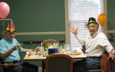 Two elderly men wearing party hats sit at a table with food, drinks, and balloons in a dining room.