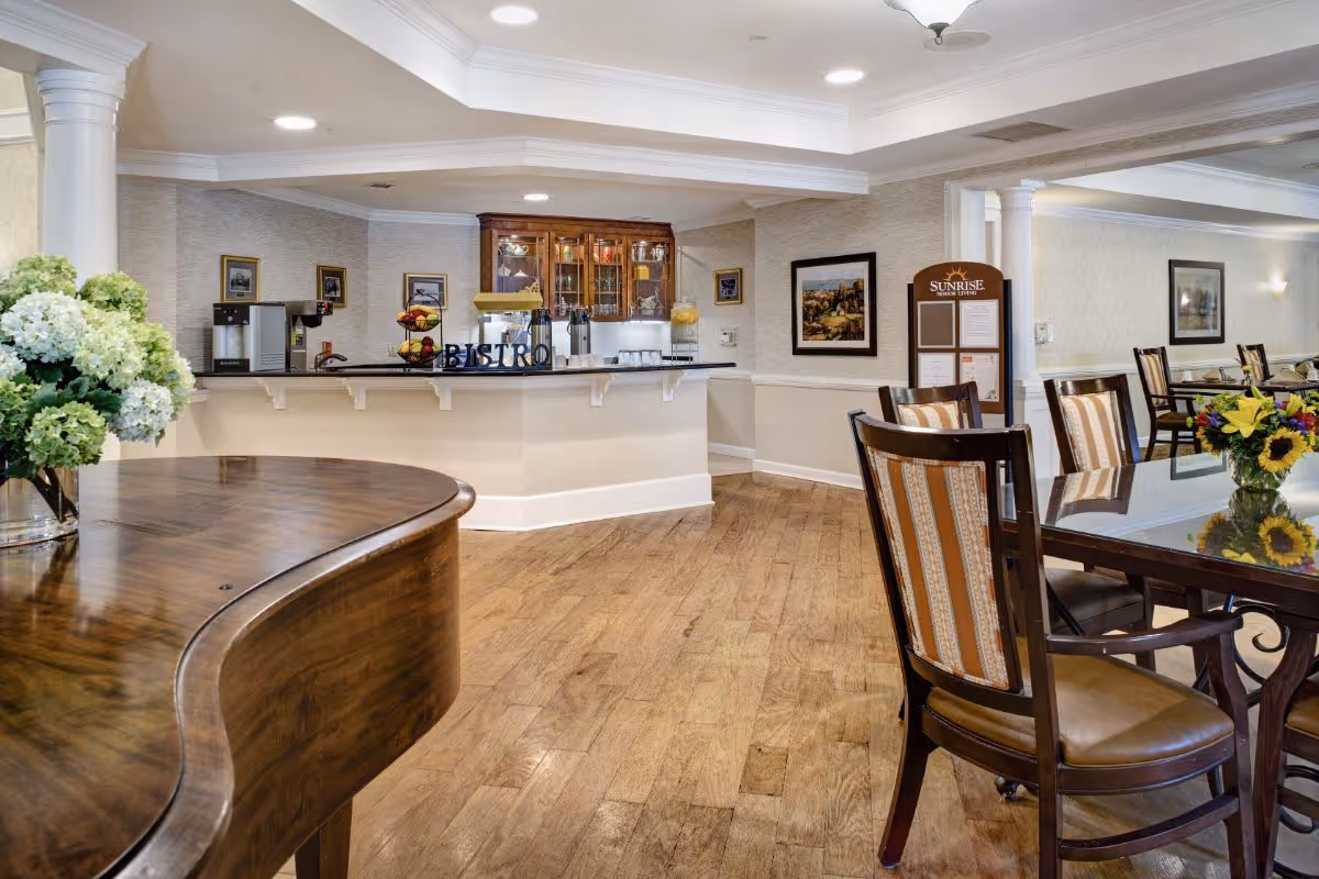 Bright communal dining area with wooden tables and chairs, a bistro counter, piano, and floral arrangements.