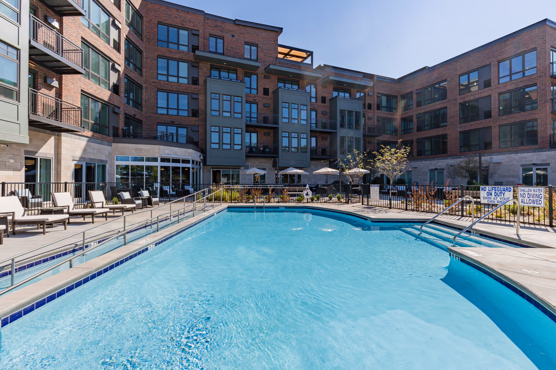 Outdoor swimming pool surrounded by lounge chairs and umbrellas, with a multi-story brick and glass building in the background under a clear blue sky.