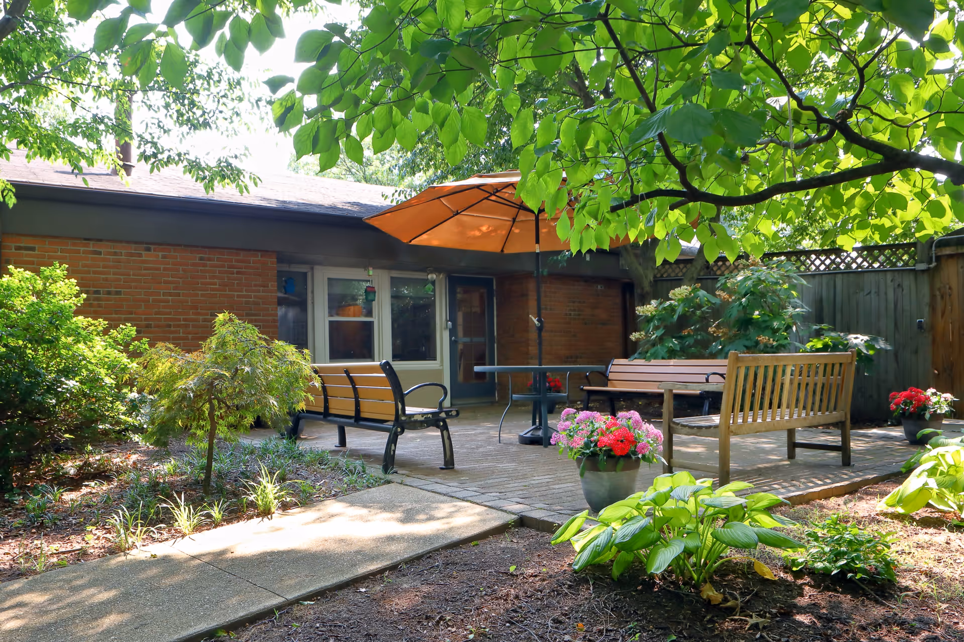 A peaceful outdoor patio area with wooden benches, a table with an orange umbrella, surrounded by green plants and flowers. The patio is adjacent to a brick building with windows and a door, and there is a wooden fence enclosing the space.
