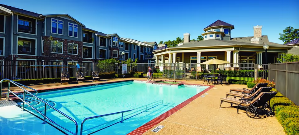 Outdoor swimming pool area at The Hamptons at Greenridge Independent Living with lounge chairs, a fenced perimeter, and a large building with multiple windows and a covered patio in the background under a clear blue sky.