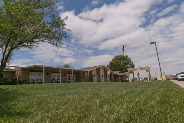 Wide view of a single-story building with a covered entrance and porch, surrounded by a well-maintained grassy lawn and a tree on the left. An American flag is flying near the entrance under a partly cloudy sky.