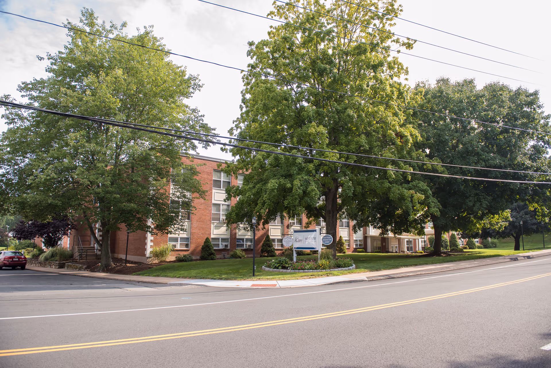 Front exterior of a brick senior living building set behind large trees with a lawn and a sign near the sidewalk.