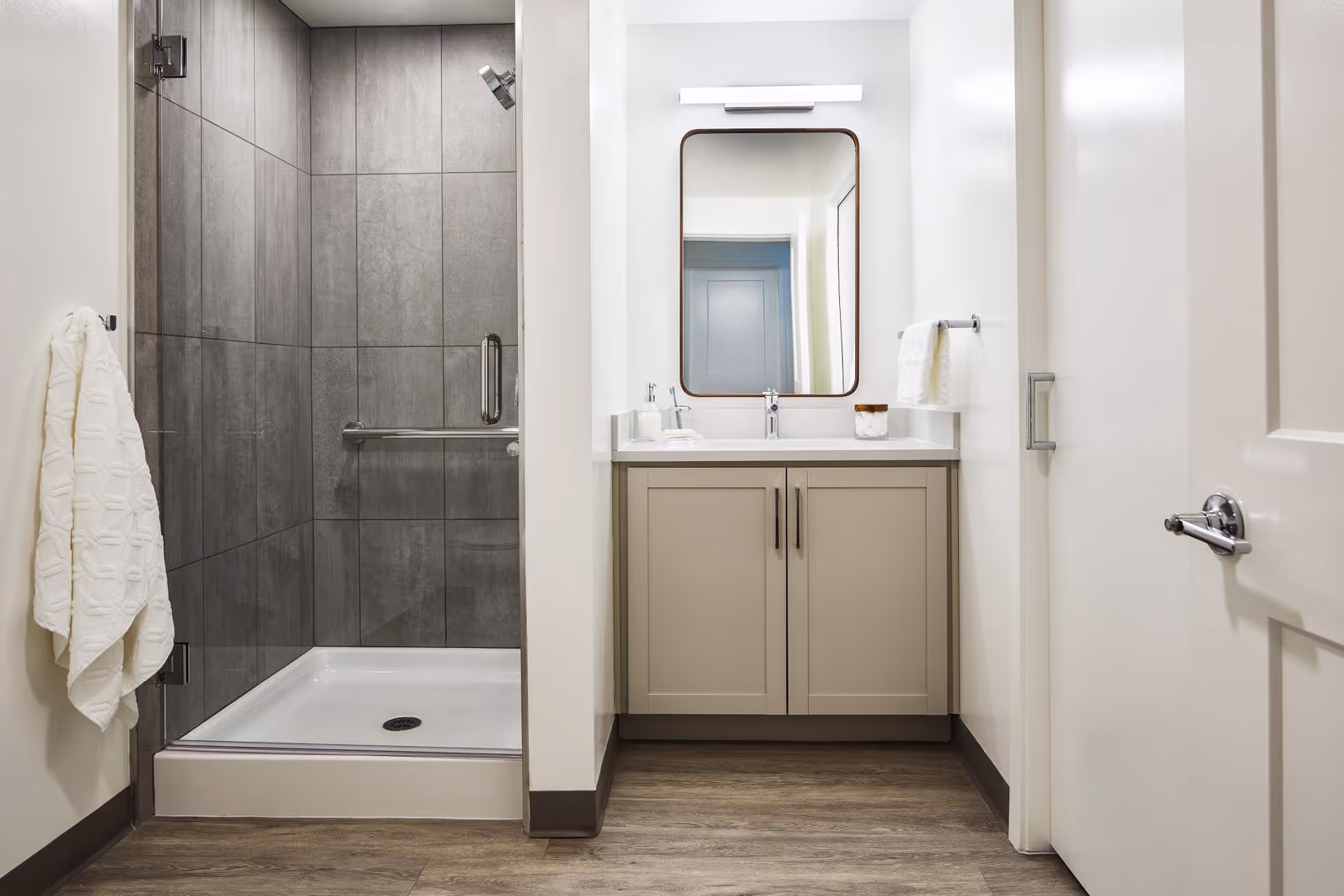 Bright modern bathroom showing a walk-in shower with gray tiles, a sink vanity with mirror, and towels.