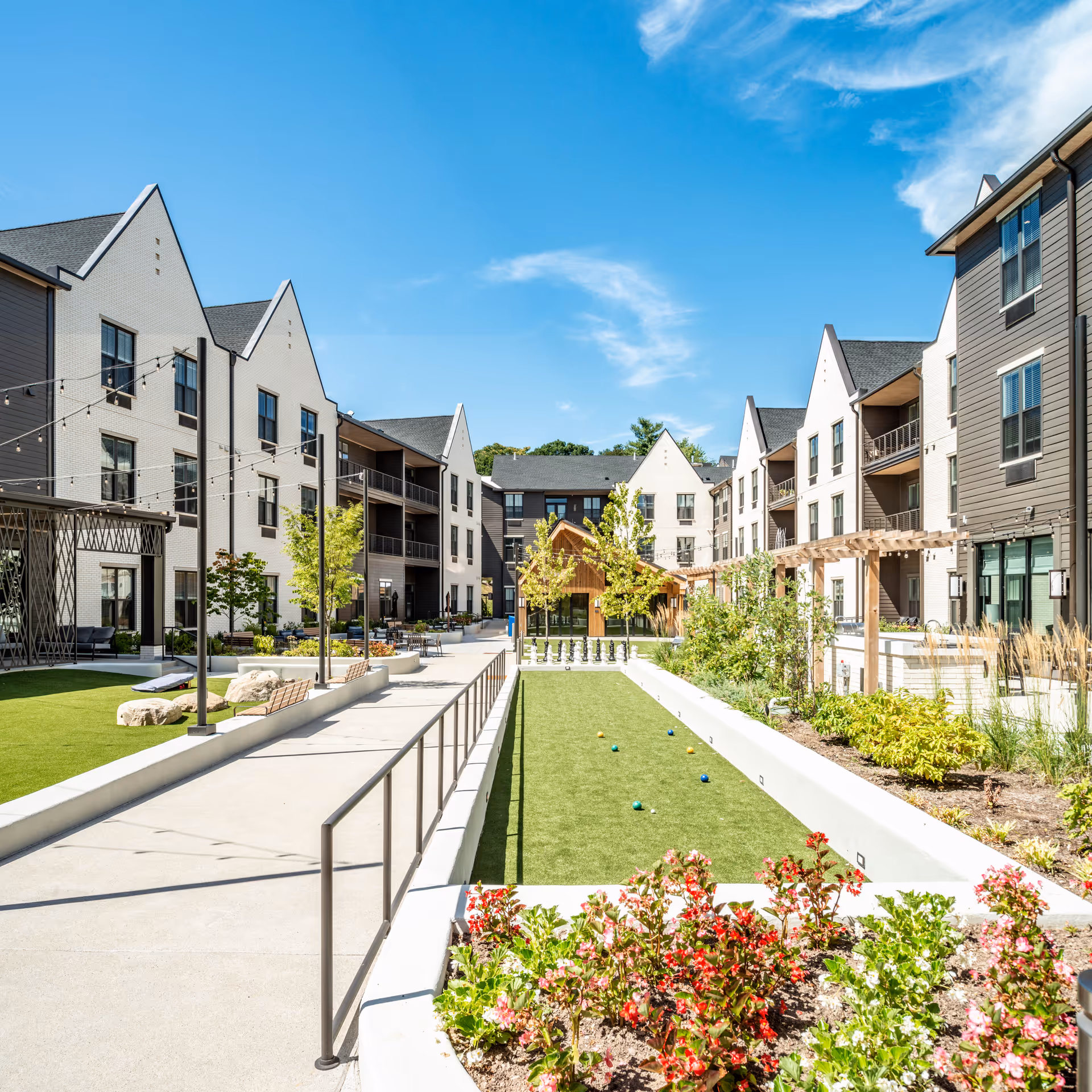Outdoor courtyard area of a senior living facility with a bocce ball court, landscaped garden beds with flowers, benches, string lights overhead, and multi-story residential buildings surrounding the space under a clear blue sky.
