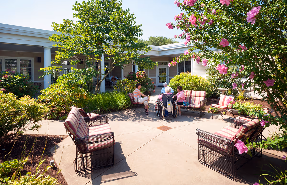 Outdoor courtyard area at Nemasket Healthcare Center with several cushioned metal chairs arranged in a circle. Four people, including one in a wheelchair, are seated and engaged in conversation. The courtyard is surrounded by greenery, flowering bushes, and trees, with a building visible in the background under a clear blue sky.