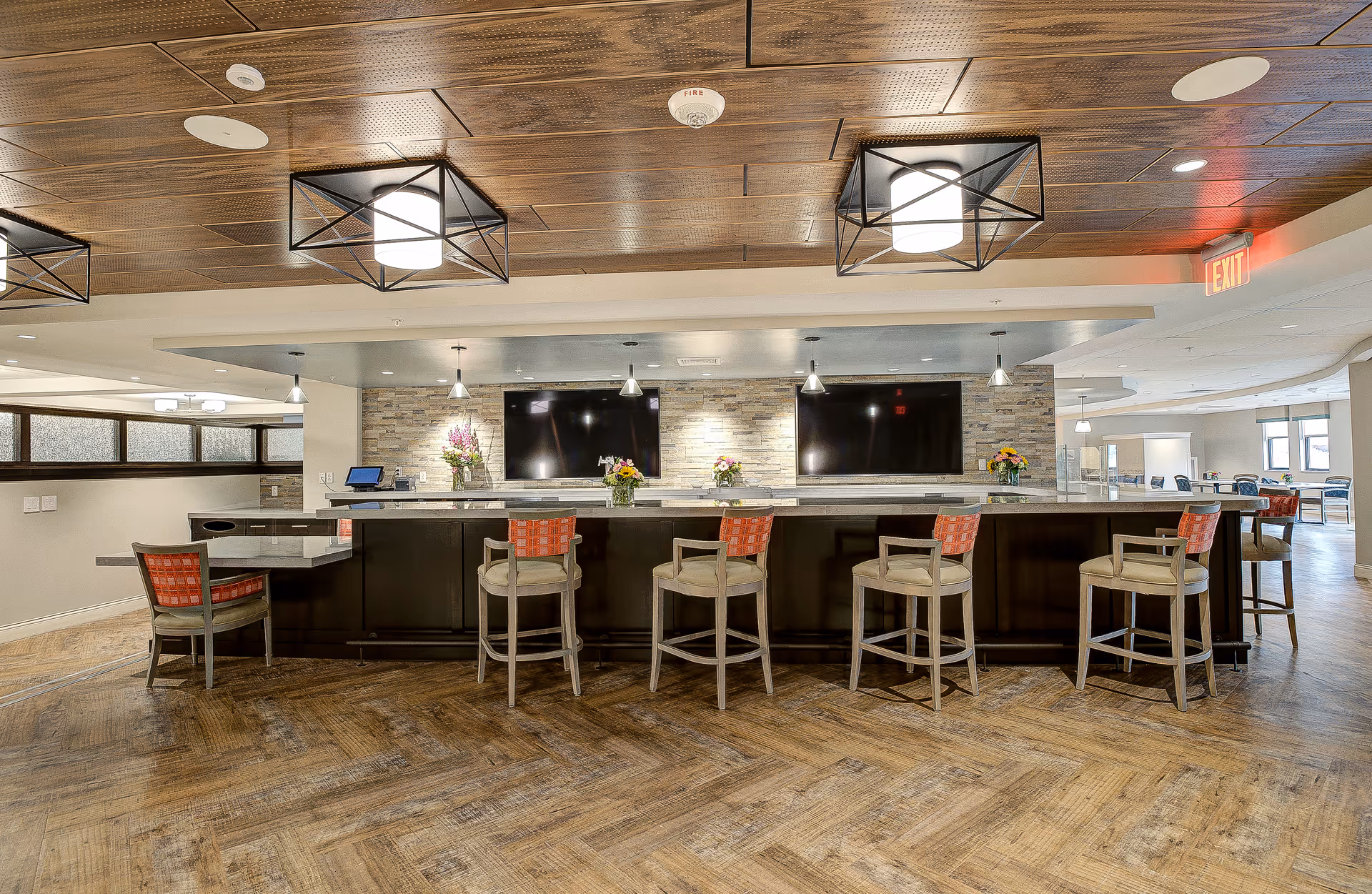 A modern common area with a long counter and six high chairs with red and beige cushions. The counter has flower arrangements and two large flat-screen TVs mounted on a stone accent wall. The ceiling features wooden panels and geometric light fixtures. The floor has a wood pattern, and there is an exit sign visible in the background.