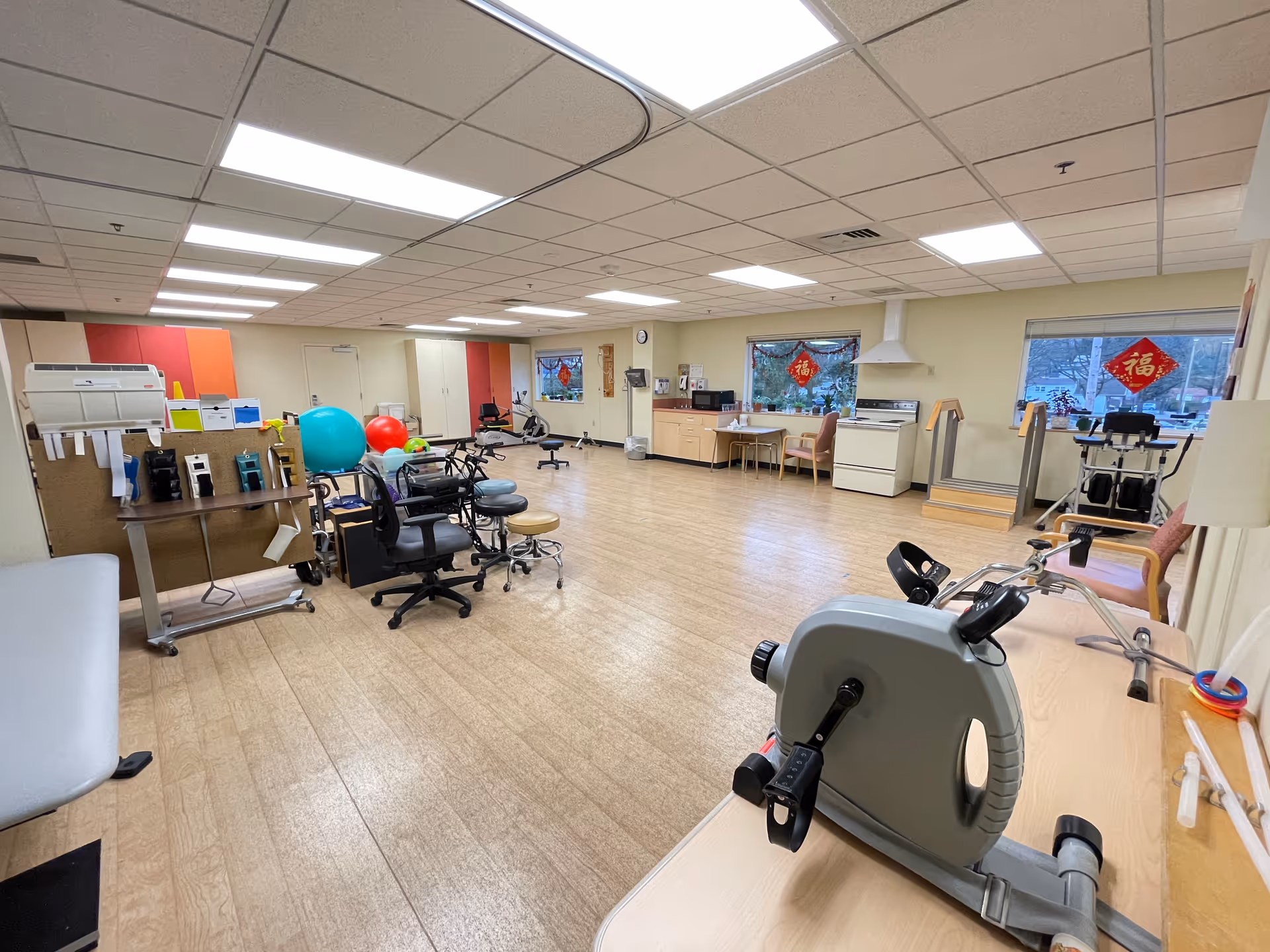A spacious room with exercise equipment including stationary bikes, therapy balls, and chairs. The room has wood flooring, fluorescent ceiling lights, and windows with red decorative hangings. There is a kitchen area with a stove, cabinets, and a sink along one wall.