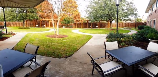 Outdoor patio area with tables and chairs under a covered section, overlooking a grassy courtyard with trees and a wooden fence in the background.