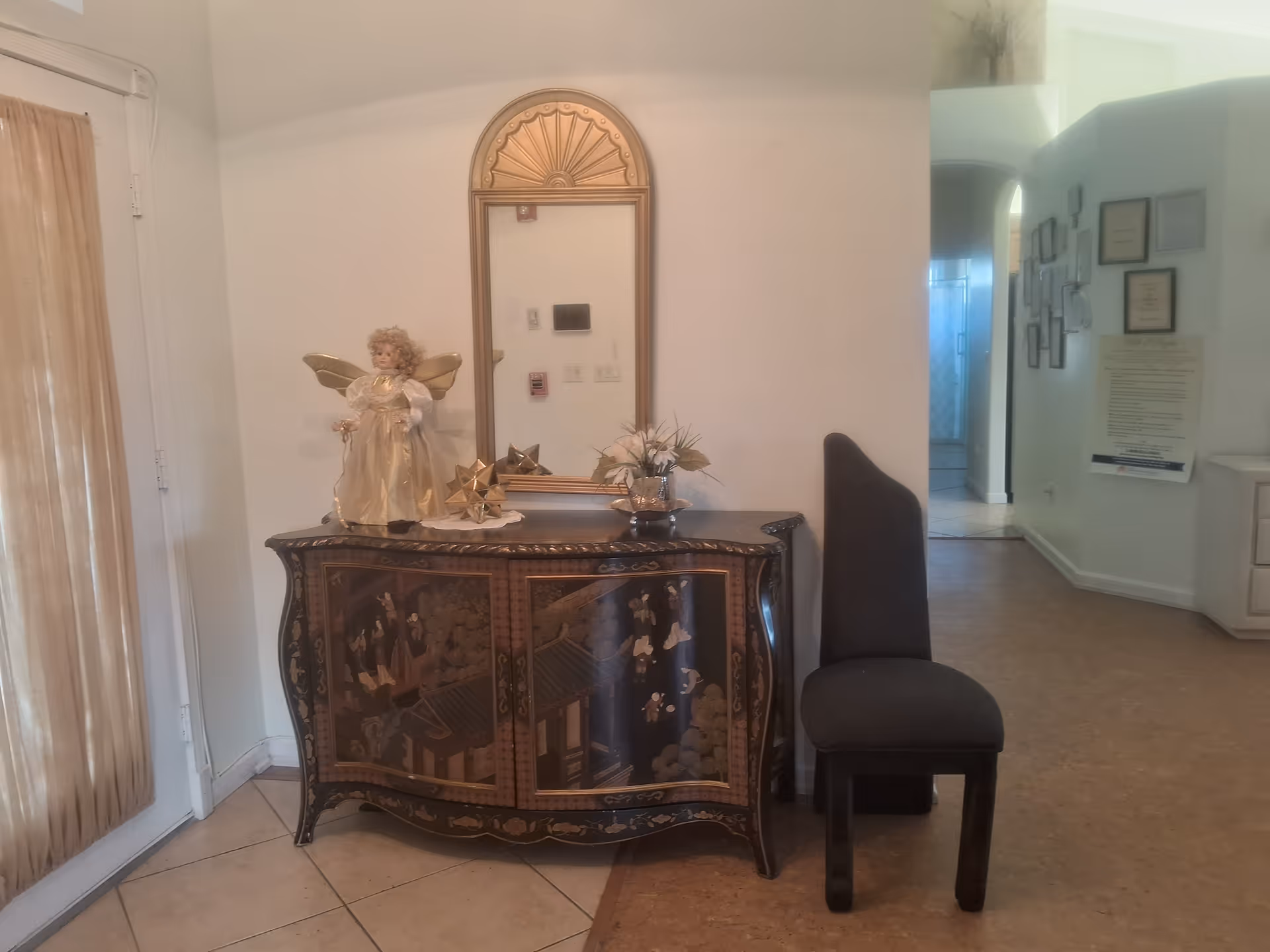 An interior corner of a room featuring a decorative wooden cabinet with Asian-inspired artwork, topped with a porcelain angel figurine, a small floral arrangement, and a golden geometric ornament. Above the cabinet is a wall-mounted mirror with a decorative golden frame. To the right of the cabinet is a tall, dark upholstered chair. The room has light-colored walls and tile flooring, with a hallway visible in the background displaying framed pictures and documents on the wall.