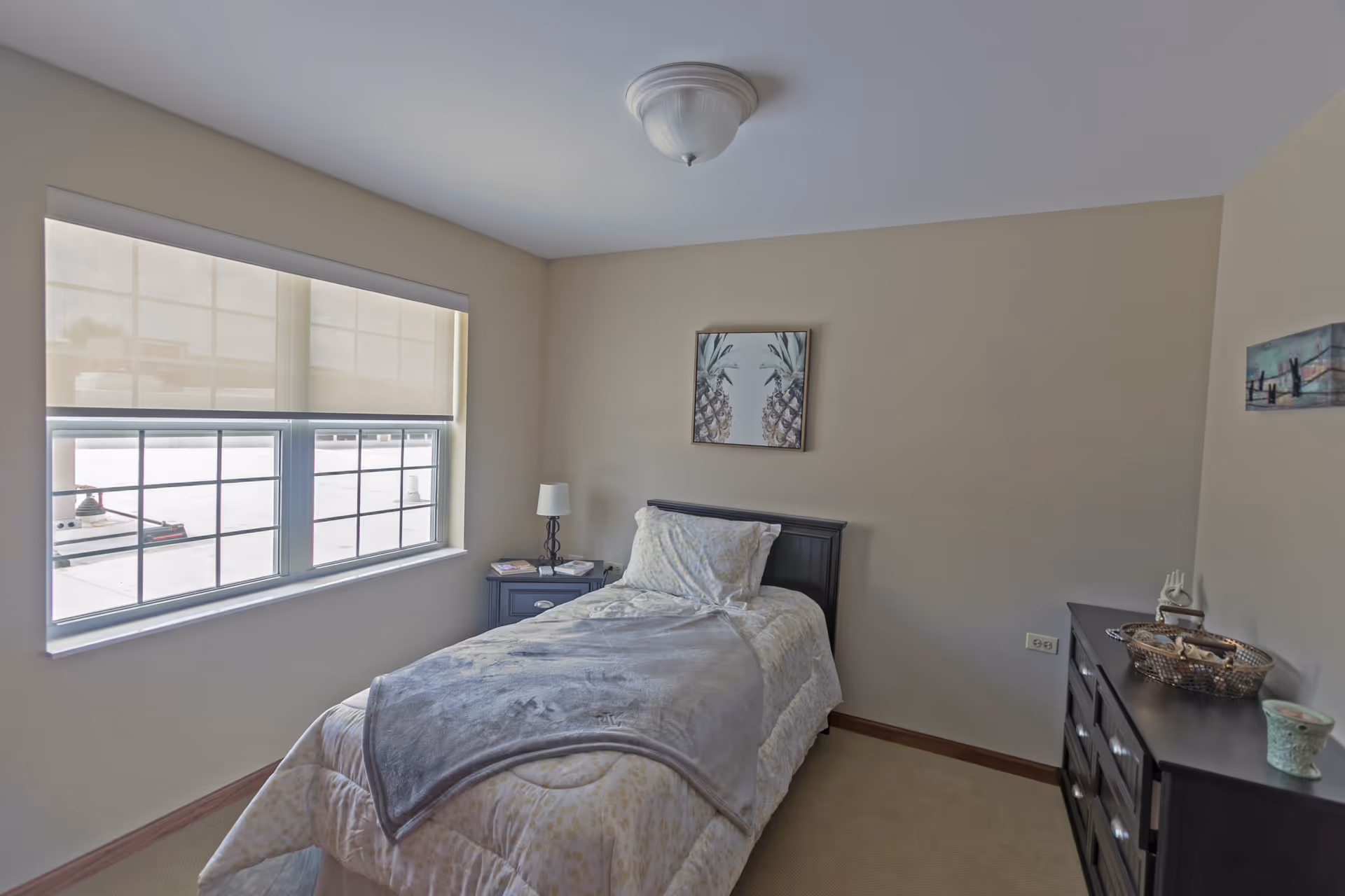 A small, neatly arranged bedroom with a single bed covered in light-colored bedding and a gray blanket. There is a nightstand with a lamp and books next to the bed, a window with a roller shade partially drawn, and a dresser with decorative items on top. The walls are painted beige and have two framed pictures hanging.