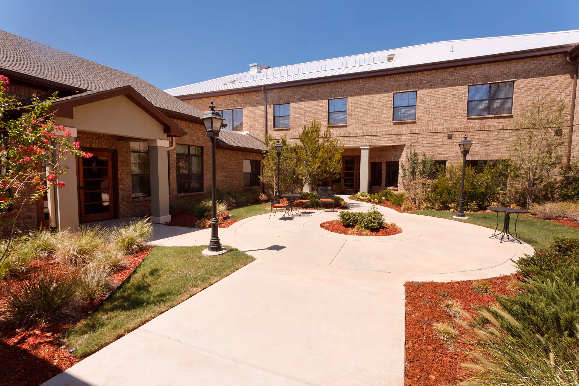 Sunlit brick courtyard with a circular paved patio, landscaping, lampposts, and outdoor seating at a senior living facility.
