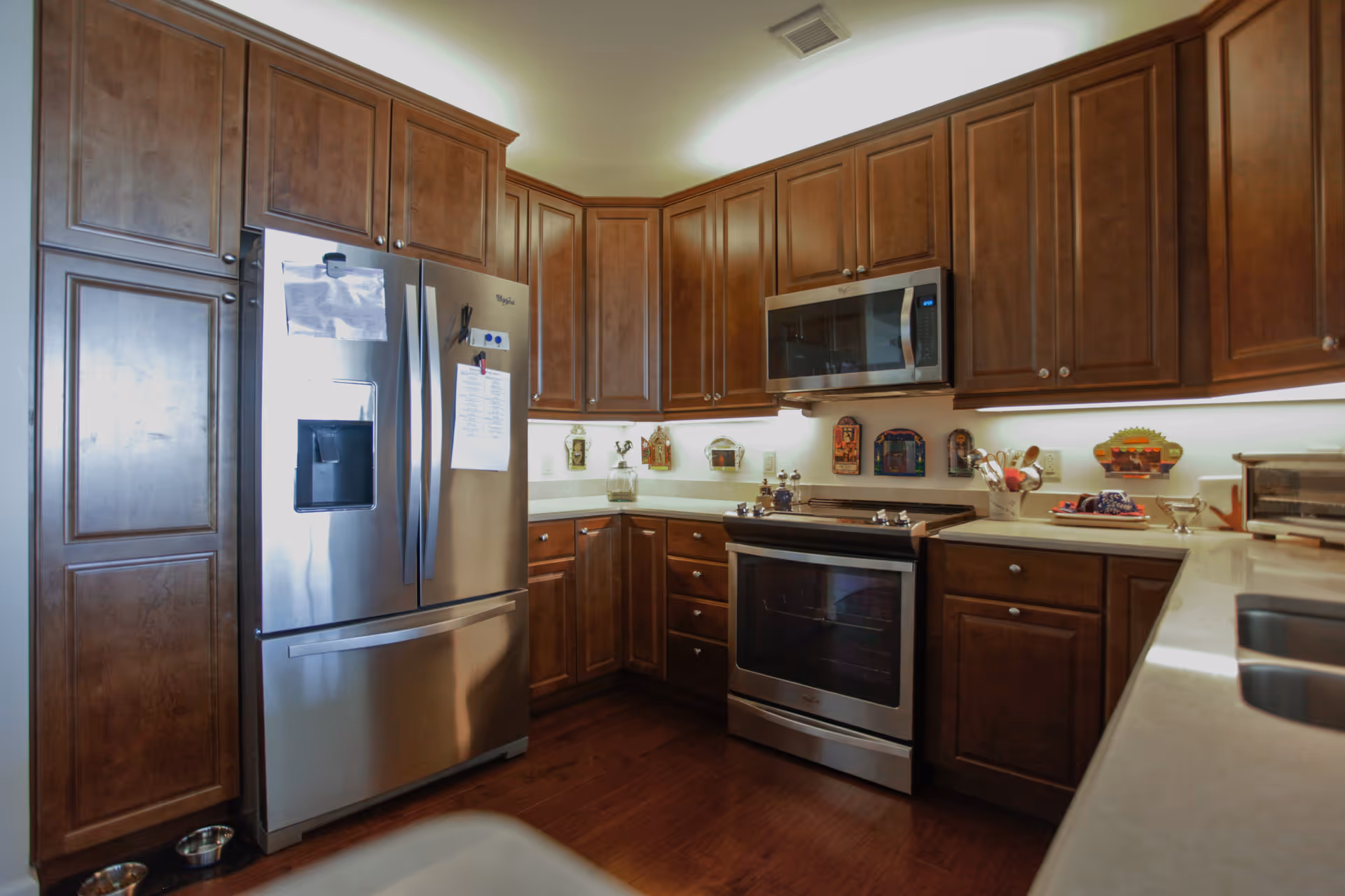 A modern kitchen with dark wooden cabinets, stainless steel refrigerator, oven, and microwave. The countertops are light-colored, and there are various decorative items and kitchen utensils on the counters.