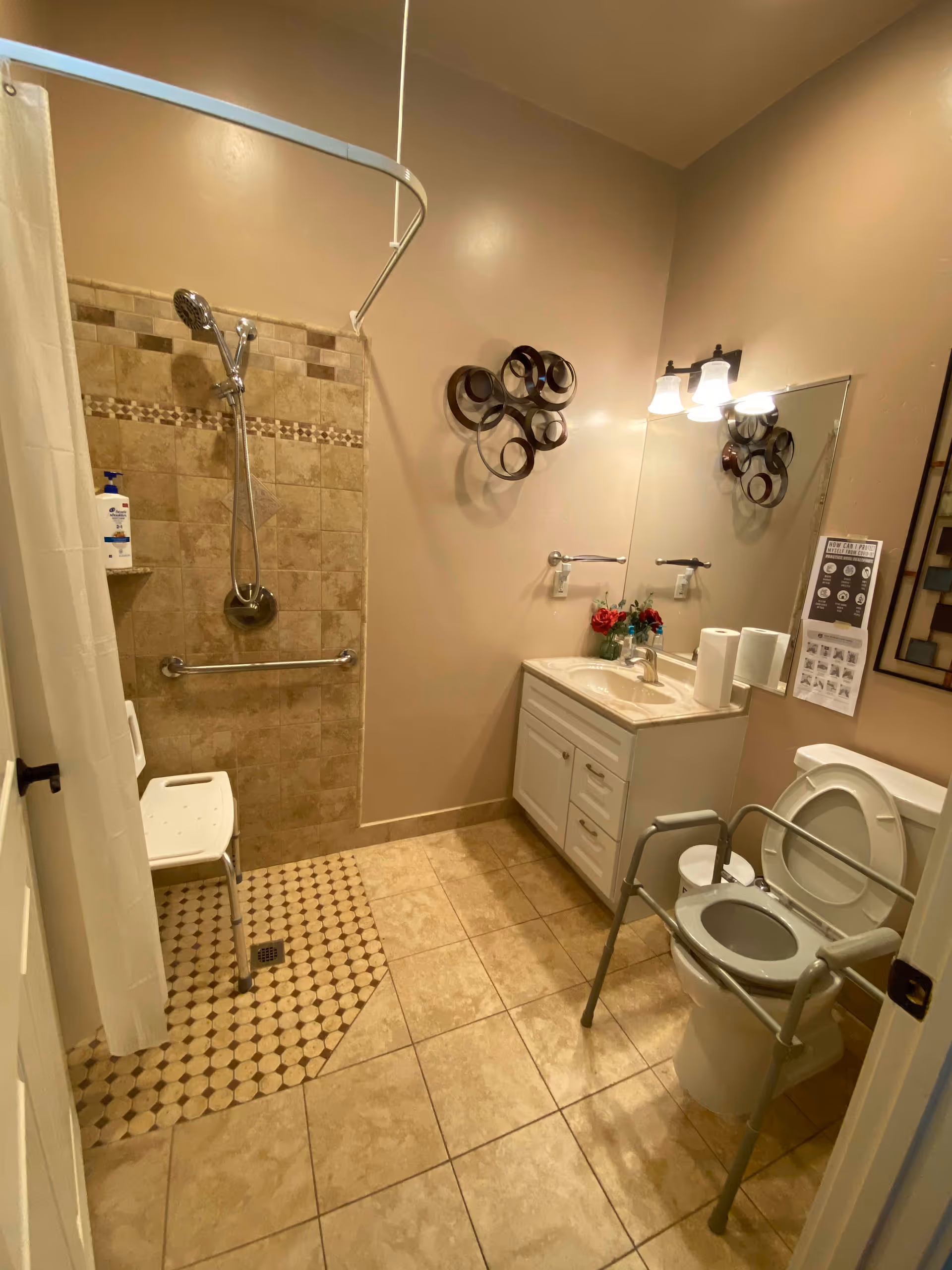 A bathroom with beige tiled floor and walls. There is a walk-in shower with a handheld showerhead, grab bar, and a white shower chair. A white vanity with a sink, mirror, and two wall-mounted lights is on the right side. Next to the vanity is a toilet with a raised seat and a metal frame for support. Decorative wall art and a small bouquet of flowers are also visible.