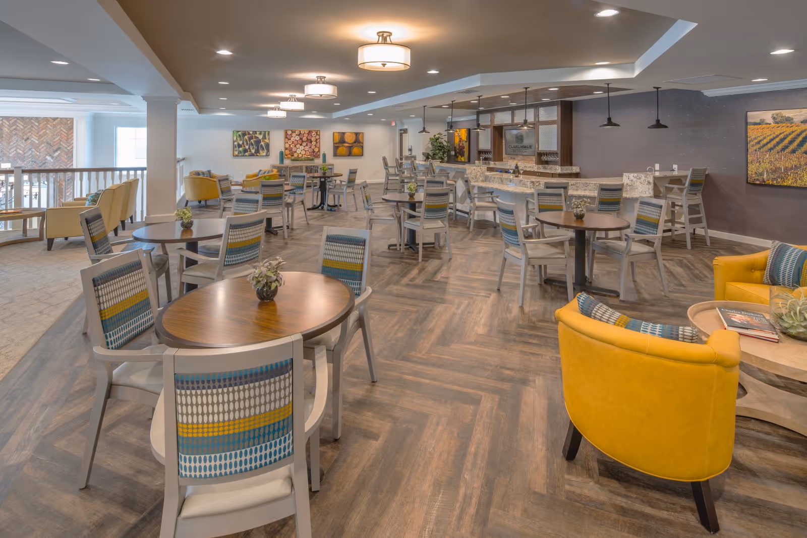 A spacious and well-lit dining area in a senior living facility featuring multiple round wooden tables with patterned chairs. The room has a modern design with a bar counter in the background, yellow armchairs, decorative paintings on the walls, and ceiling lights providing warm illumination.