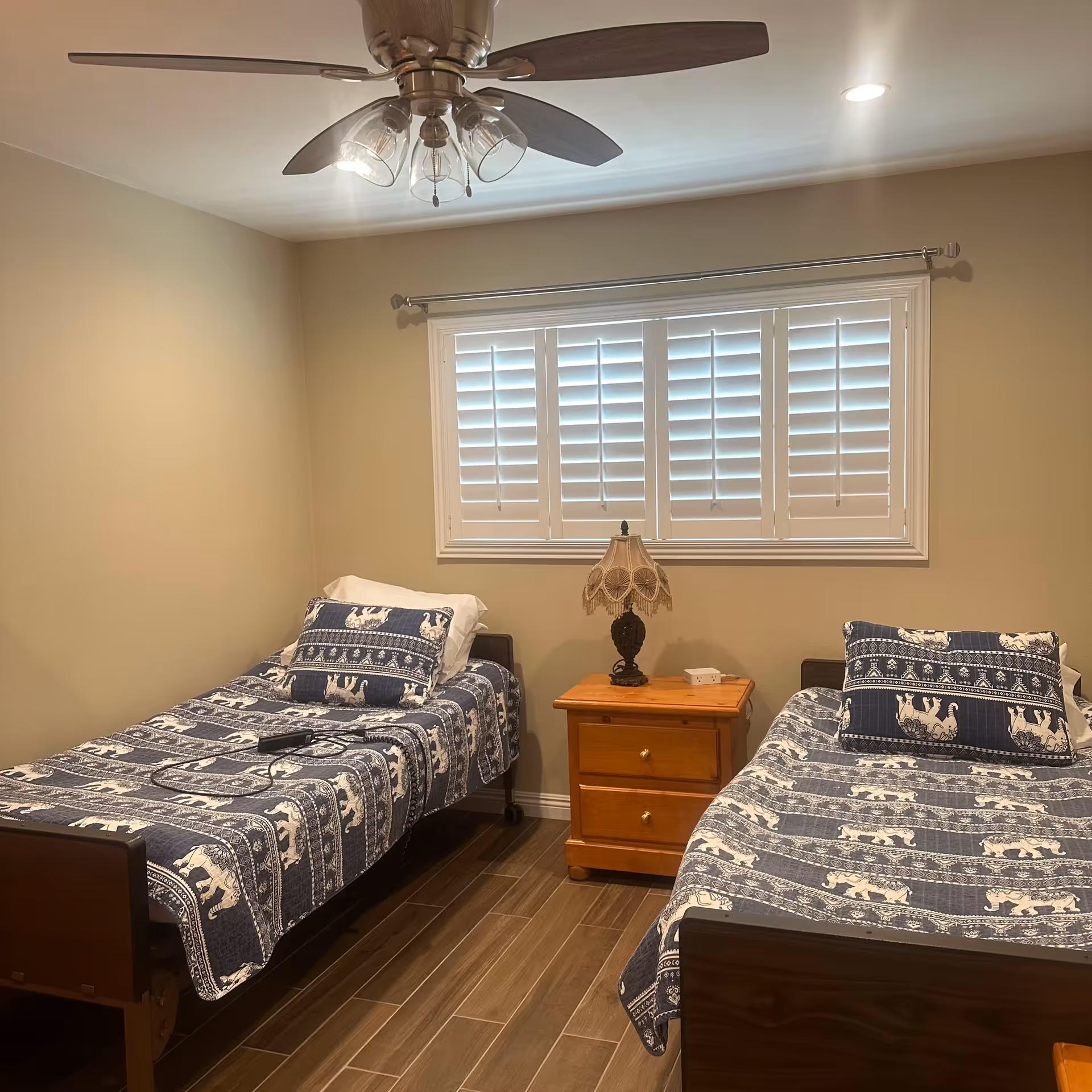 A small bedroom with two twin beds dressed in blue patterned quilts, a wooden nightstand and lamp between them, and a window with white plantation shutters.