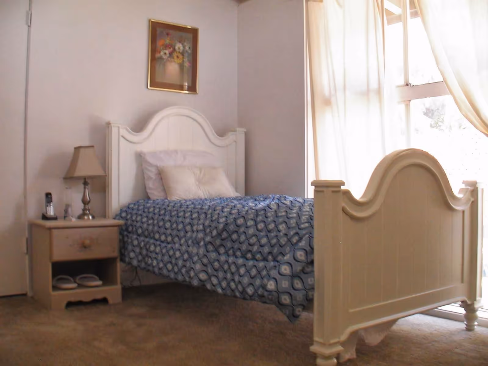 A bright bedroom with a single white wooden bed, blue patterned bedspread, nightstand with a lamp and phone, and a window with curtains.