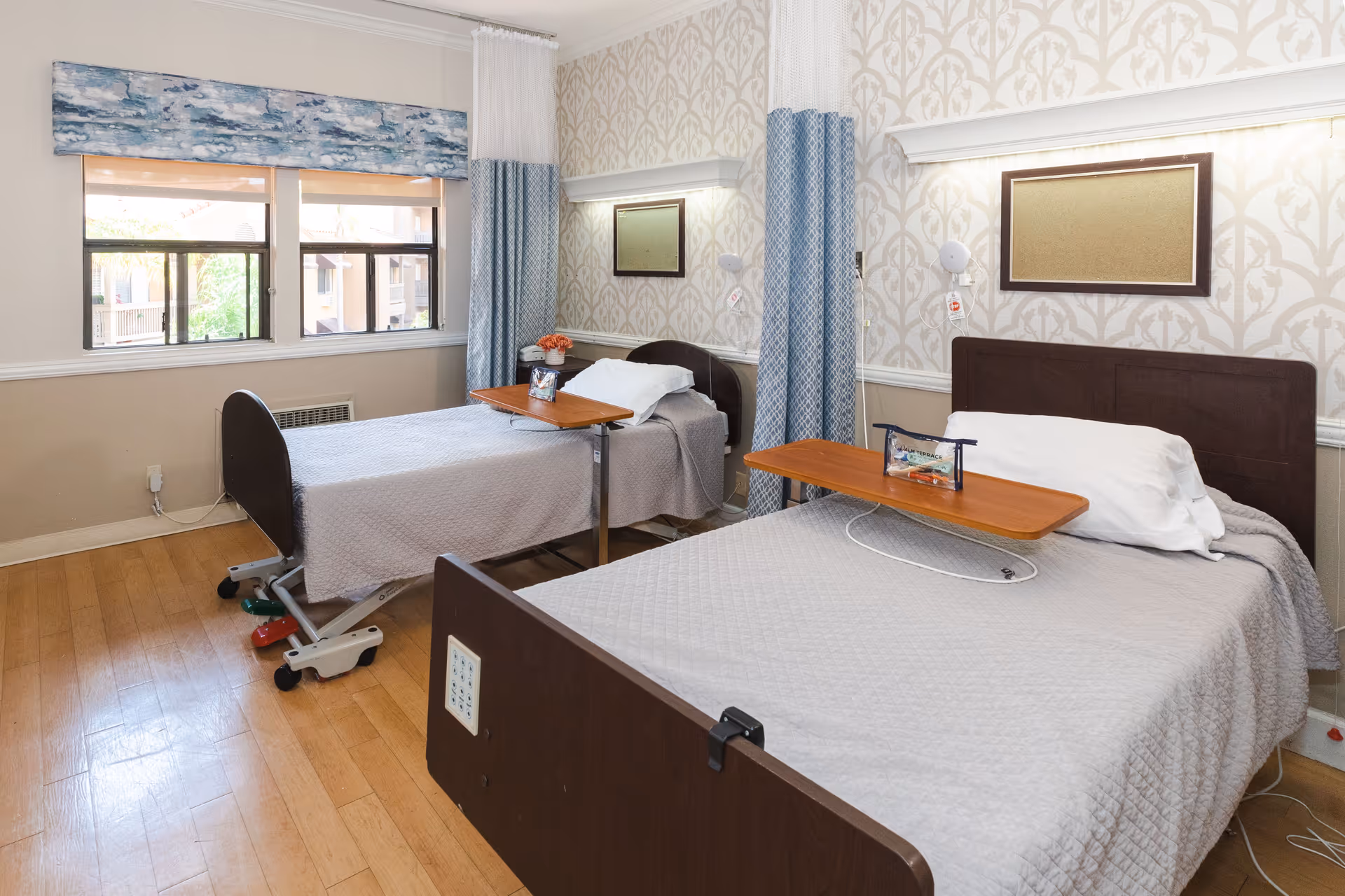 A healthcare facility room with two hospital beds, each with a wooden overbed table, white pillows, and gray bedspreads. The room has light-colored patterned wallpaper, two windows with blue and white curtains, and wooden flooring. There are wall-mounted reading lights and a framed corkboard above each bed.