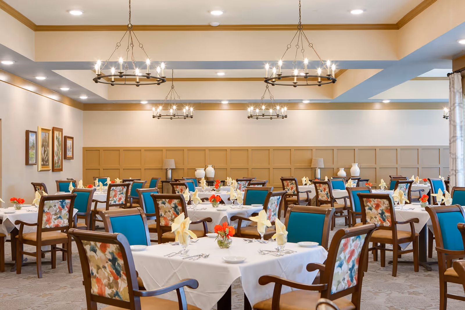 A spacious dining room with multiple tables covered in white tablecloths, each set with folded yellow napkins, glassware, and silverware. The chairs have wooden frames with teal and floral upholstery. The room features beige walls with wooden paneling, decorative vases on side tables, framed artwork, and several round chandeliers hanging from the ceiling providing warm lighting.