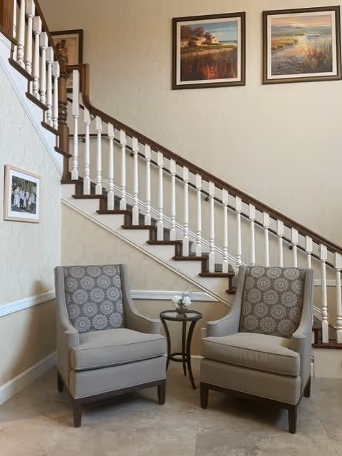 Two gray upholstered armchairs with patterned cushions are placed side by side in front of a staircase with white balusters and a wooden handrail. A small round table with a flower vase is positioned between the chairs. The wall behind features two framed landscape paintings and a smaller framed photo near the bottom of the stairs.