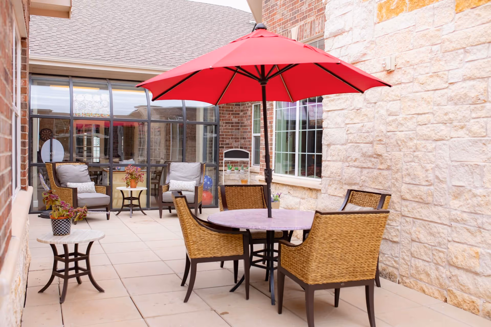 Outdoor patio area with a round table and four wicker chairs under a large red umbrella. There are two cushioned armchairs with a small table between them against a brick and stone wall. Potted plants are placed on the tables and the patio floor is tiled.