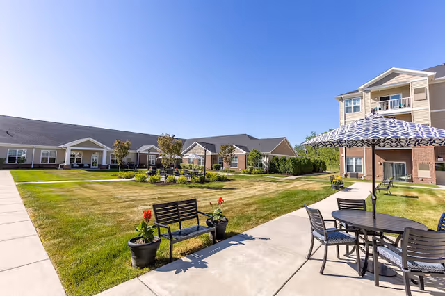 Sunny landscaped courtyard with lawn, benches, a patio table and umbrella, and surrounding apartment-style buildings.