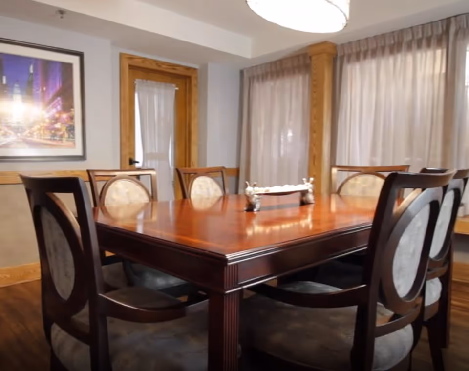 A dining room with a polished wooden table surrounded by six cushioned chairs. The room features wooden trim around the door and windows, with sheer curtains covering the windows. A framed picture hangs on the wall, and a decorative centerpiece is placed on the table.