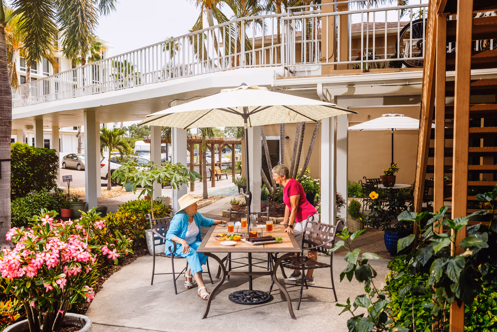 Two elderly women enjoying a conversation at an outdoor patio table with an umbrella, surrounded by lush greenery and flowers at a senior living facility.