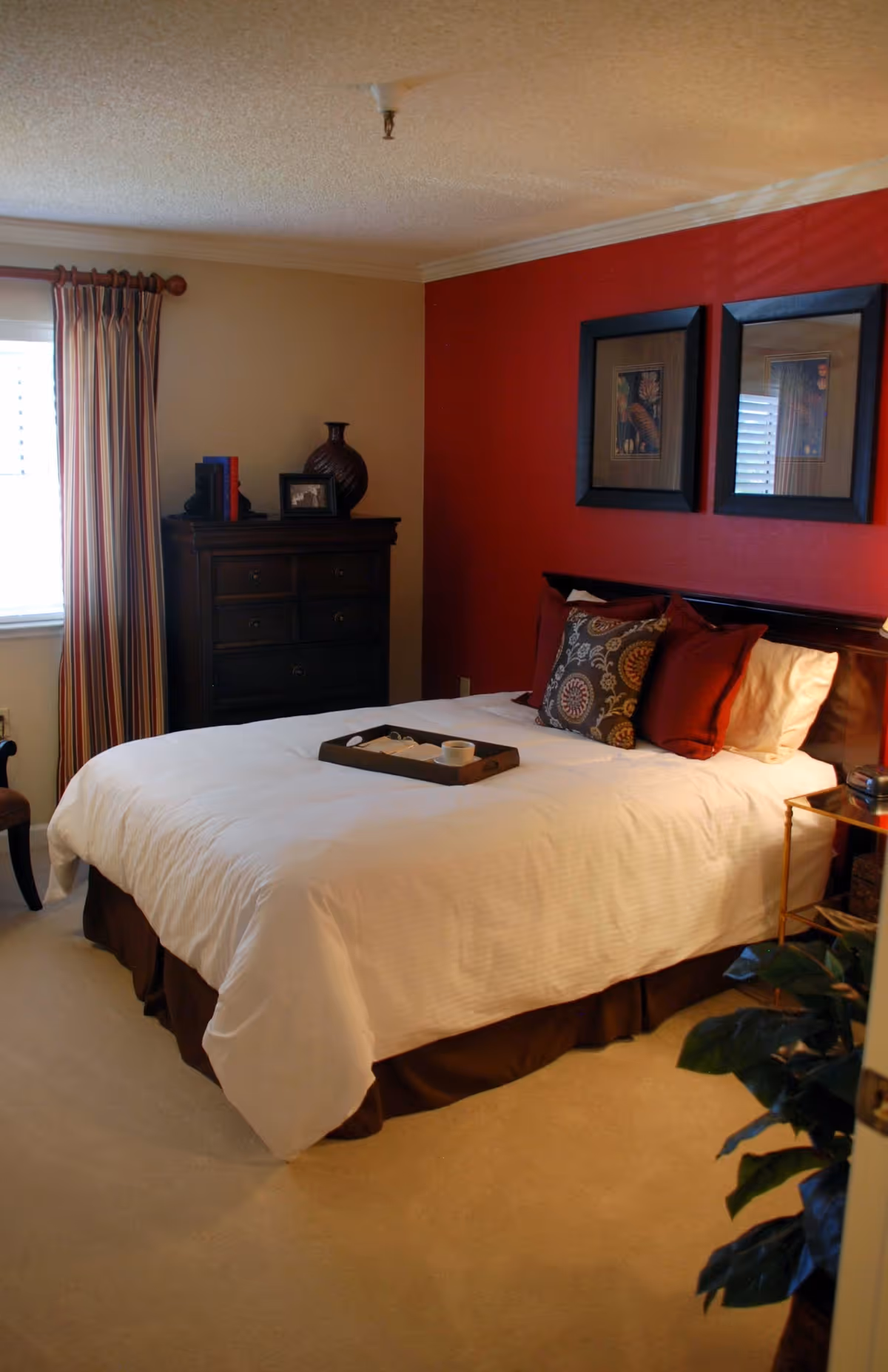 A cozy bedroom with a white bedspread and decorative pillows on a bed against a red accent wall. Two framed pictures hang above the bed. A dark wooden dresser with decorative items and books is positioned near a window with striped curtains. A small side table and a leafy plant are also visible.