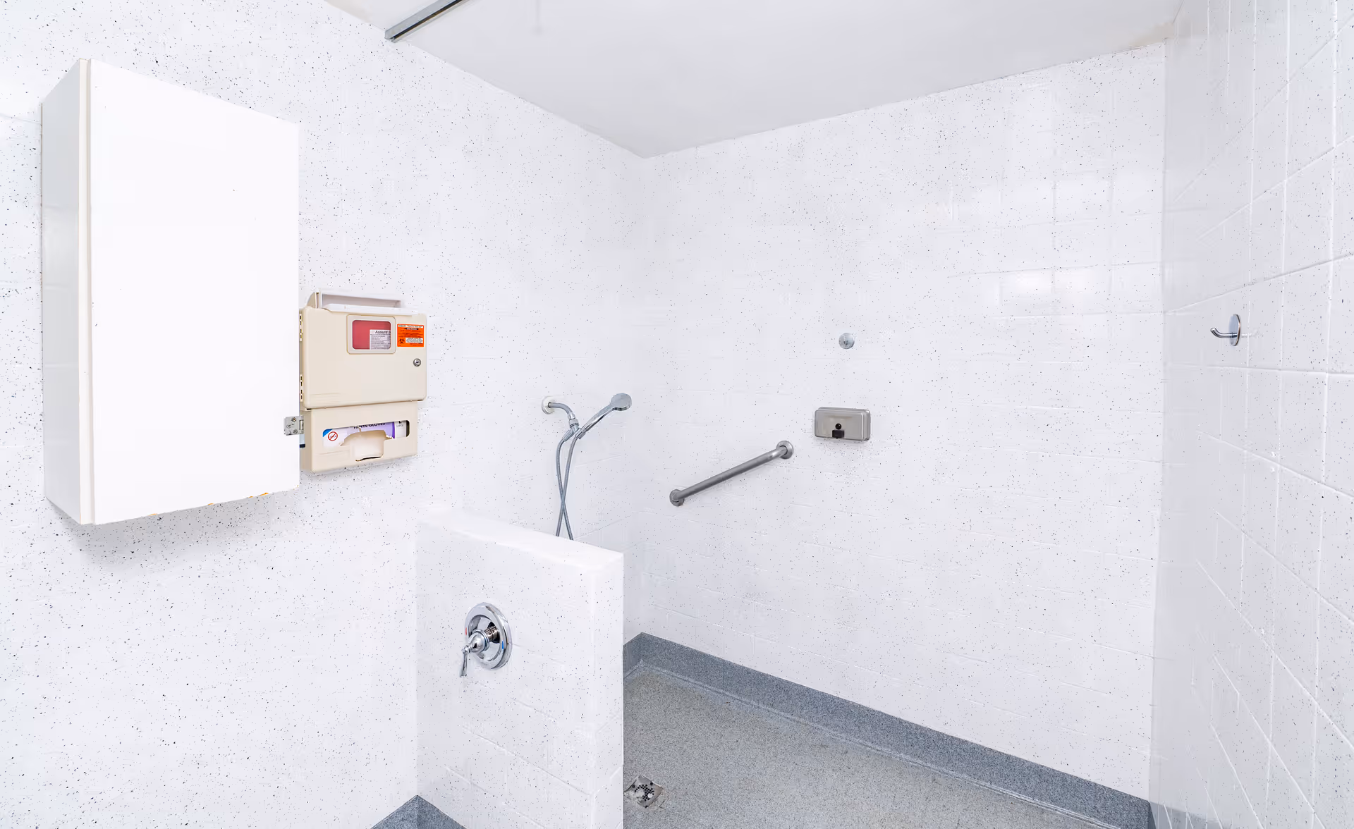 A clean, white tiled shower room with a handheld showerhead, a grab bar on the wall, a soap dispenser, and a small half wall with a faucet. The floor is gray and there is a white cabinet and control panel mounted on the wall.