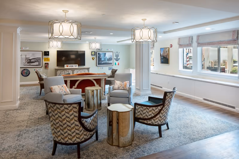 A bright and spacious living room area in a senior living facility with four armchairs arranged around two small gold-colored side tables. The room features large windows with patterned Roman shades, a flat-screen TV mounted on the wall, and various decorative wall art including Italian-themed signs and pictures. The floor is carpeted with a patterned rug, and there are modern ceiling light fixtures.