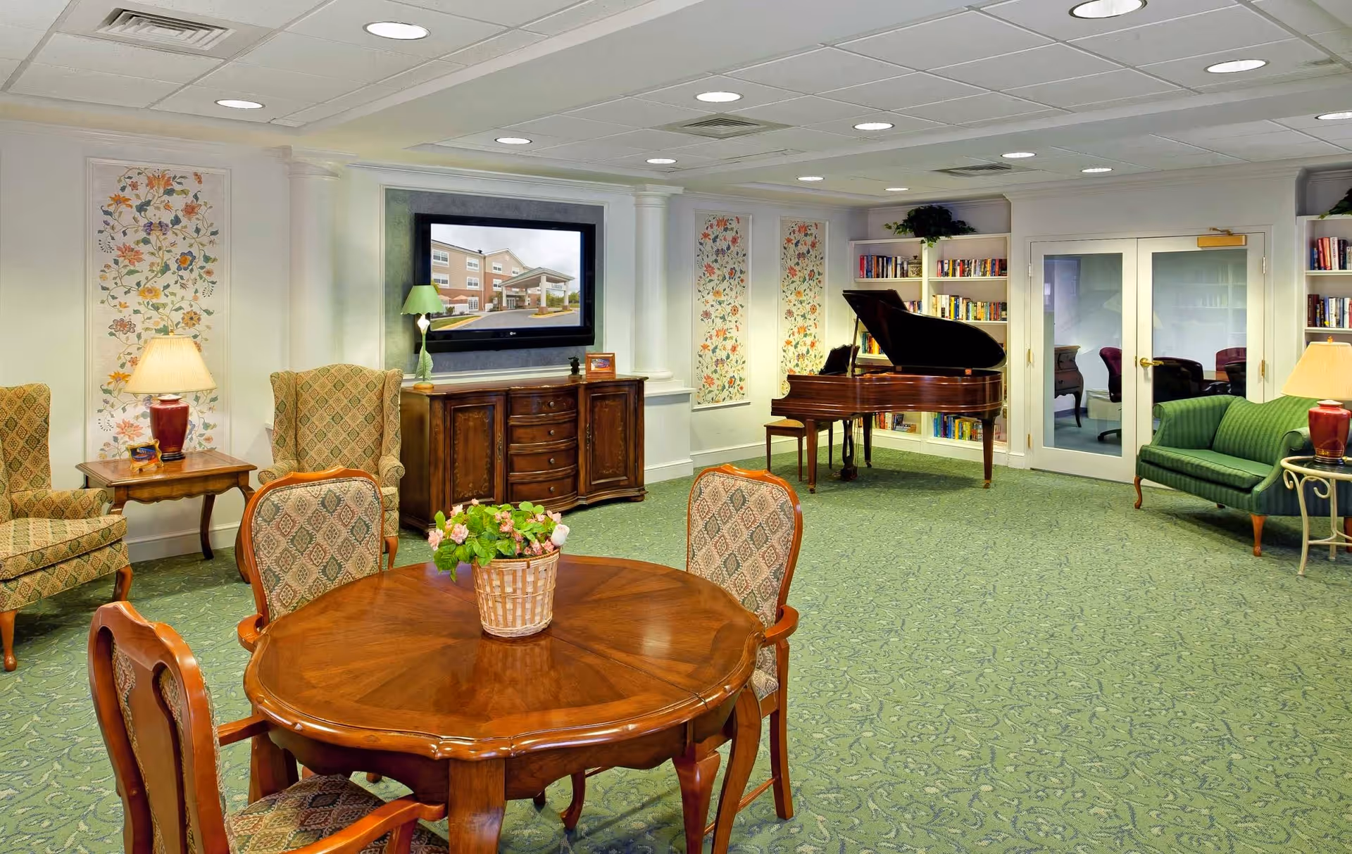 A cozy living room area with green patterned carpet, a round wooden table with four upholstered chairs, two armchairs, a green sofa, a grand piano, bookshelves filled with books, a wooden cabinet with a flat-screen TV mounted above it, and decorative floral wall panels. The room is well-lit with ceiling lights and table lamps.