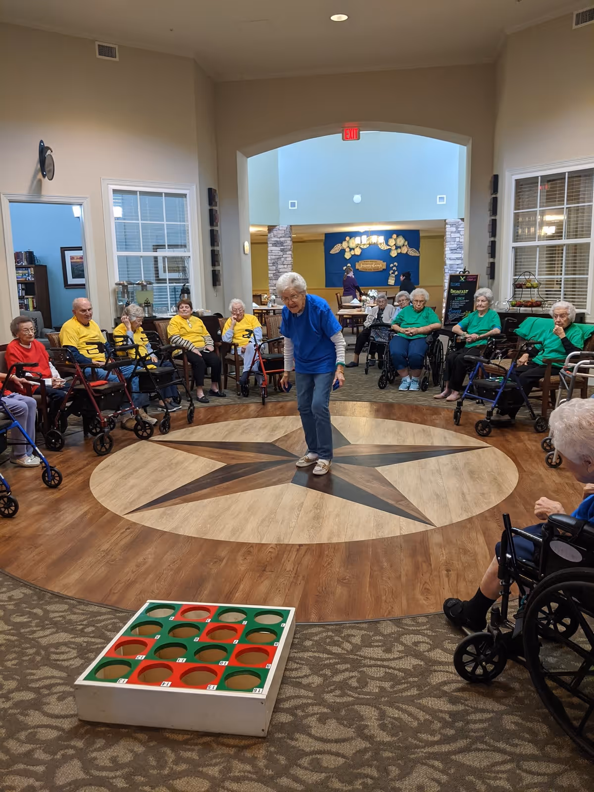 A group of elderly people sitting in a circle in a common area of a senior living facility. Most are seated in wheelchairs or using walkers. One elderly woman in a blue shirt is standing in the center of a circular wooden floor with a star design, appearing to participate in a game. A large game board with red and green holes is on the carpeted floor in the foreground. The room has high ceilings and large windows.