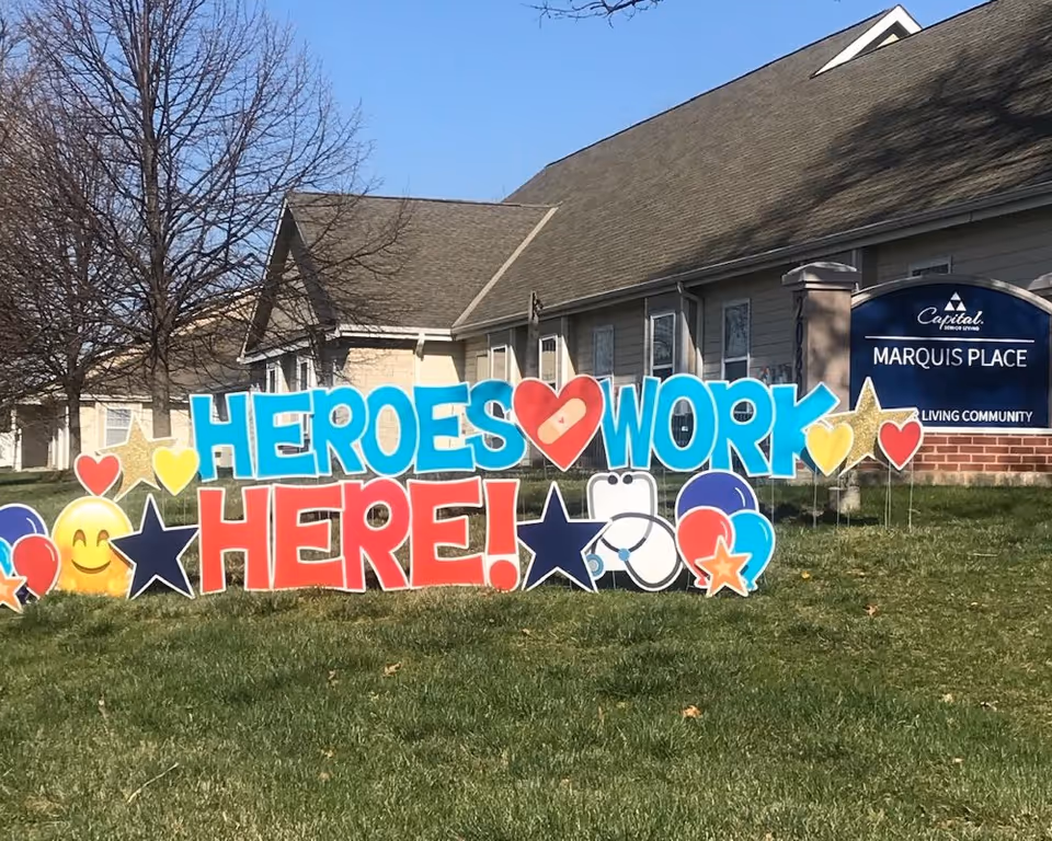 Outdoor view of Marquis Place senior living community with a colorful yard sign that reads 'HEROES WORK HERE!' surrounded by stars, hearts, balloons, and a smiley face emoji. The building and a sign for Marquis Place are visible in the background under a clear blue sky.